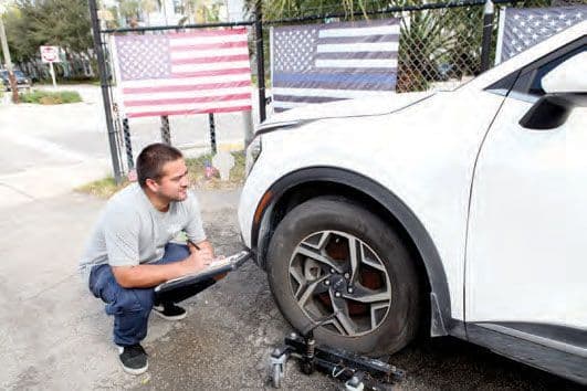 Shop technician crouched beside a white SUV writing an estimate on a clipboard